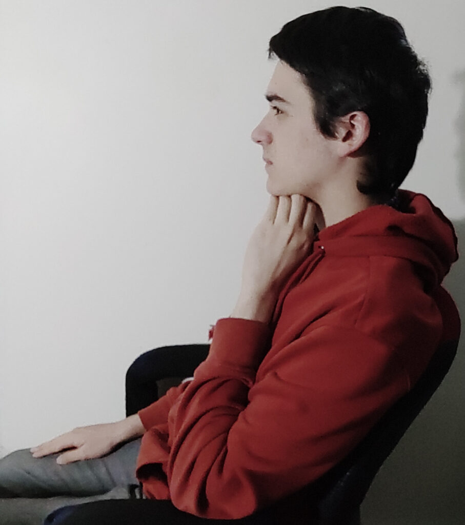 Cette image présente Phil avec un habit brun-rouge et des patalons gris. Il est assis, a sa main droite sur sa cuisse, a la tête droite et met sa main en dessous de sa tête pour la maintenir.